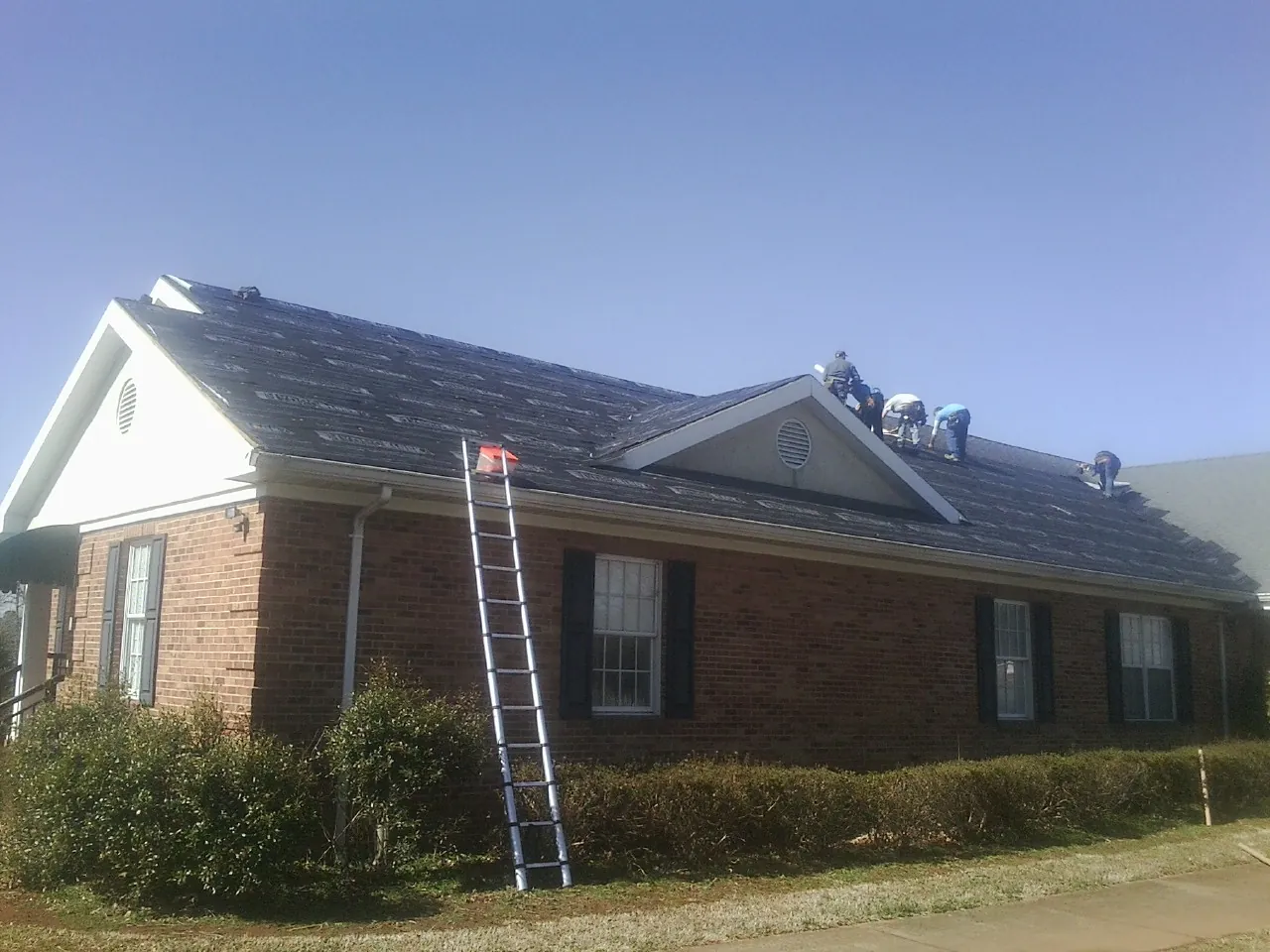 Crew tearing shingles off a brick ranch roof with a ladder set — Metal Roof Repair work by our crew in Osteen